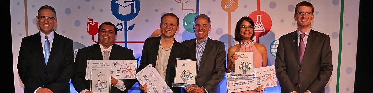 6 pessoas em frente a um painel colorido, como imagens que remetem a educação, ciência, pesquisa entre outros. Os 4 do meio seguram seus prêmios e cheques.