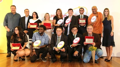 14 pessoas no palco posando para a foto. As pessoas formam duas fileiras no palco. Em pé há alguns vencedores com seus prêmios, além do André Araujo (presidente da Shell) e a realizadora do evento. Embaixo, há 5 vencedores agachados com seus prêmios.