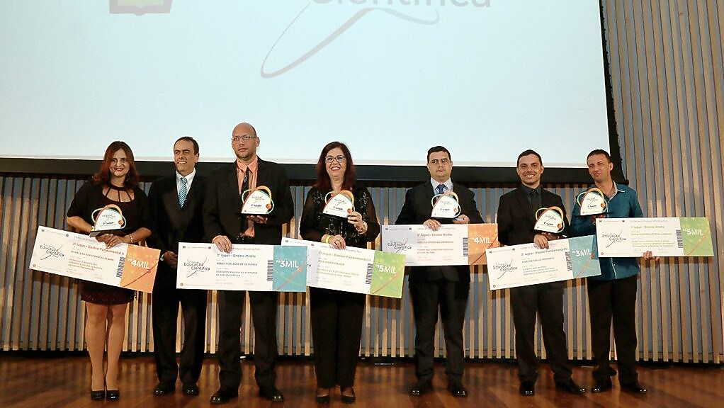 6 vencedores do Prêmio de Educação Científica no palco, segurando seus prêmios e cheques. Da direita para a esquerda, se encontra o presidente da Shell, André Araujo. Ao fundo há um telão com a logo da Shell e escrito terceiro prêmio de educação científica