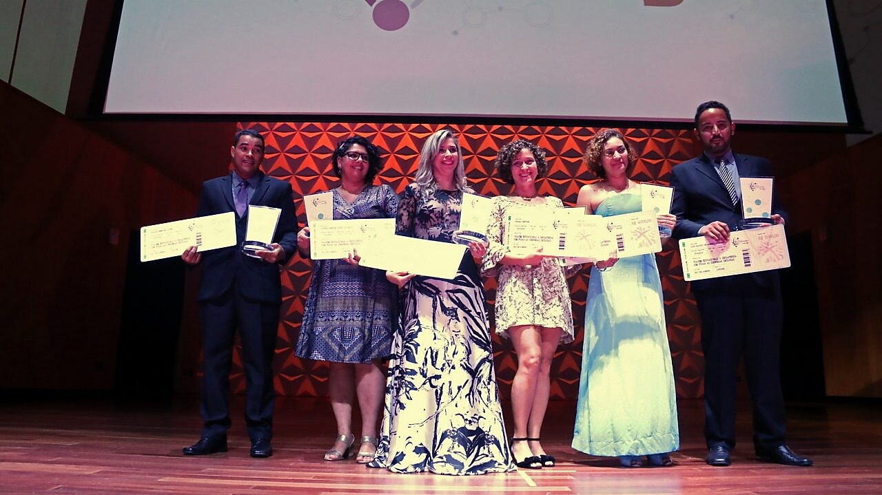 6 vencedores no palco do Prêmio de Educação Científica, segurando seus cheques e prêmios. Ao fundo há um telão com a logo da BG e do prêmio.