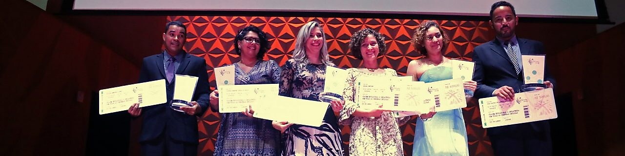 6 vencedores no palco do Prêmio de Educação Científica, segurando seus cheques e prêmios. Ao fundo há um telão com a logo da BG e do prêmio.