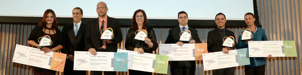 6 vencedores do Prêmio de Educação Científica no palco, segurando seus prêmios e cheques. Da direita para a esquerda, se encontra o presidente da Shell, André Araujo. Ao fundo há um telão com a logo da Shell e escrito terceiro prêmio de educação científica