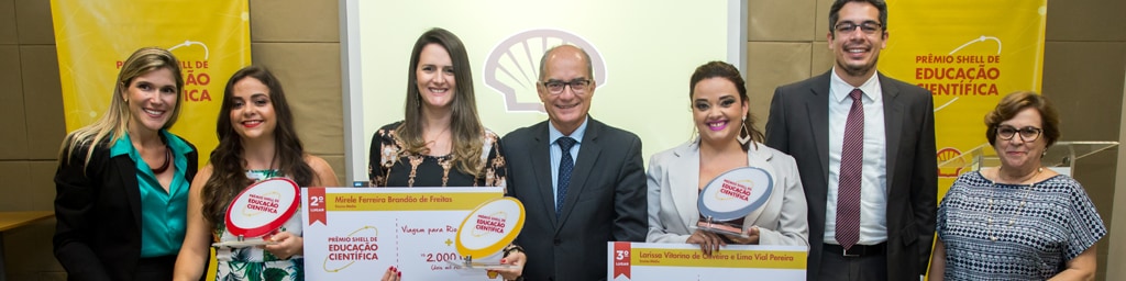 7 pessoas em pé posando para a foto. Dentre elas, 3 seguram o prêmio e o cheque de primeiro, segundo e terceiro lugar. Ao fundo há dois banners escrito Prêmio Shell de Educação Científica e um telão pequeno com a logo da Shell, a concha.