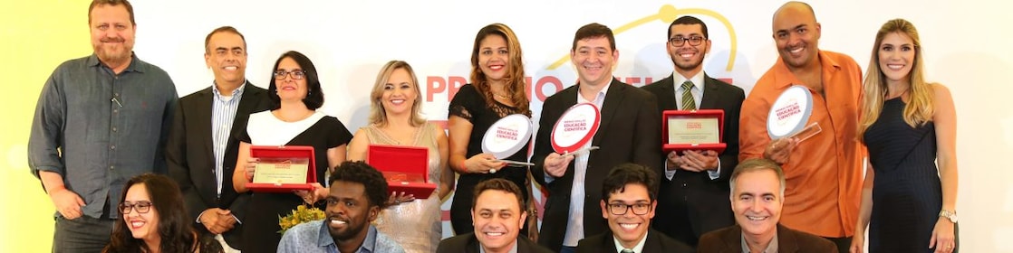14 pessoas no palco posando para a foto. As pessoas formam duas fileiras no palco. Em pé há alguns vencedores com seus prêmios, além do André Araujo (presidente da Shell) e a realizadora do evento. Embaixo, há 5 vencedores agachados com seus prêmios.