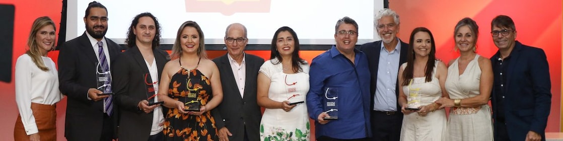11 pessoas em pé posando para a foto. Dentre eles há 5 vencedores do Prêmio Shell de Educação Científica do Espírito Santo com seu trofeu, a apresentadora do prêmio e alguns dos avaliadores. Ao fundo temos uma luz avermelhada e um painel com a logo da concha.