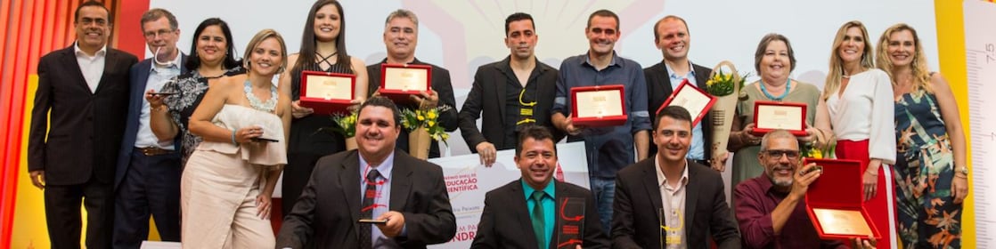 16 pessoas no palco posando para a foto. As pessoas formam duas fileiras no palco. Em pé atrás com há alguns vencedores com seus prêmio, flores e um cheque. Além de duas pessoas da Shell e o presidente, André Araujo. Embaixo, há 4 pessoas agachados com seus cheques e prêmios. Ao fundo vemos painel amarelado e a concha da Shell.