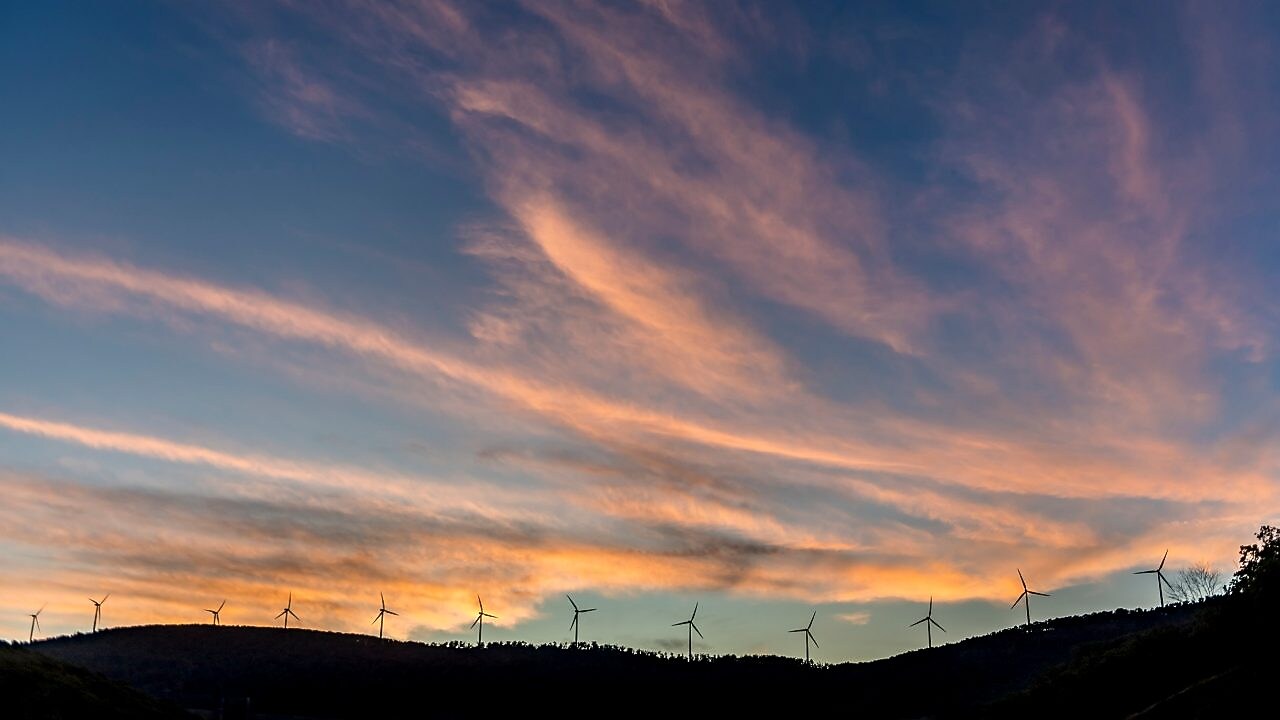 Imagem distante de um monte com turbinas eólicas lado a lado, ao entardecer. Vemos um céu azul escuro e nuvens alaranjadas.