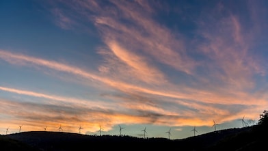 Imagem distante de um monte com turbinas eólicas lado a lado, ao entardecer. Vemos um céu azul escuro e nuvens alaranjadas.