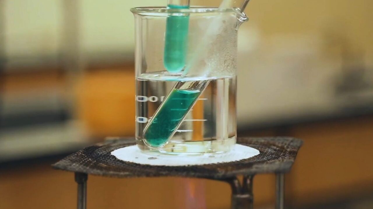 Imagem de experimento em laboratório. Um béquer de vidro transparente com liquido incolor, aquecendo um tubo de ensaio com um líquido verde translúcido.