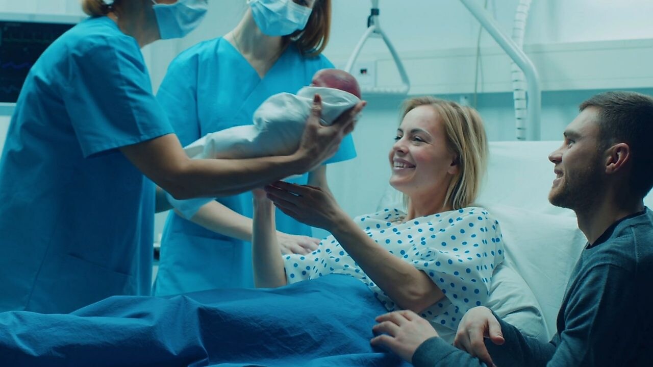 No quarto de um hospital, vemos duas profissionais da saúde, uma delas entrega um bebê recém-nascido a uma mulher que está ao lado de um homem.