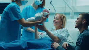 No quarto de um hospital, vemos duas profissionais da saúde, uma delas entrega um bebê recém-nascido a uma mulher que está ao lado de um homem.