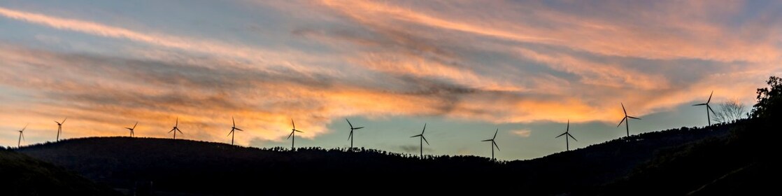 Imagem distante de um monte com turbinas eólicas lado a lado, ao entardecer. Vemos um céu azul escuro e nuvens alaranjadas.