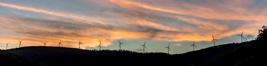 Imagem distante de um monte com turbinas eólicas lado a lado, ao entardecer. Vemos um céu azul escuro e nuvens alaranjadas.