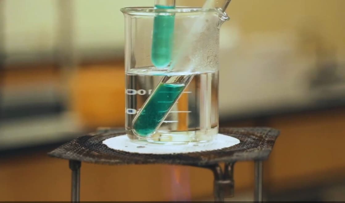 Imagem de experimento em laboratório. Um béquer de vidro transparente com liquido incolor, aquecendo um tubo de ensaio com um líquido verde translúcido.