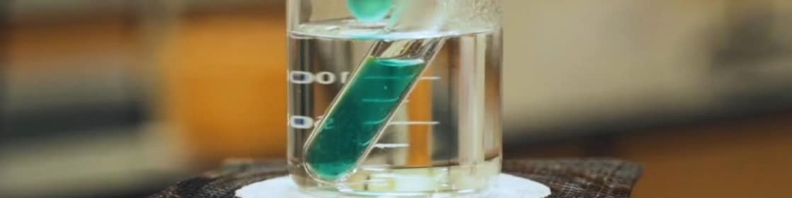 Imagem de experimento em laboratório. Um béquer de vidro transparente com liquido incolor, aquecendo um tubo de ensaio com um líquido verde translúcido.
