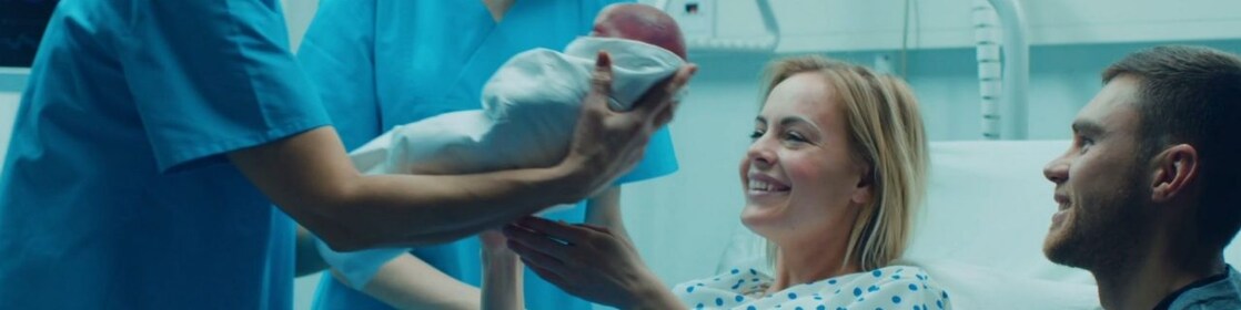 No quarto de um hospital, vemos duas profissionais da saúde, uma delas entrega um bebê recém-nascido a uma mulher que está ao lado de um homem.