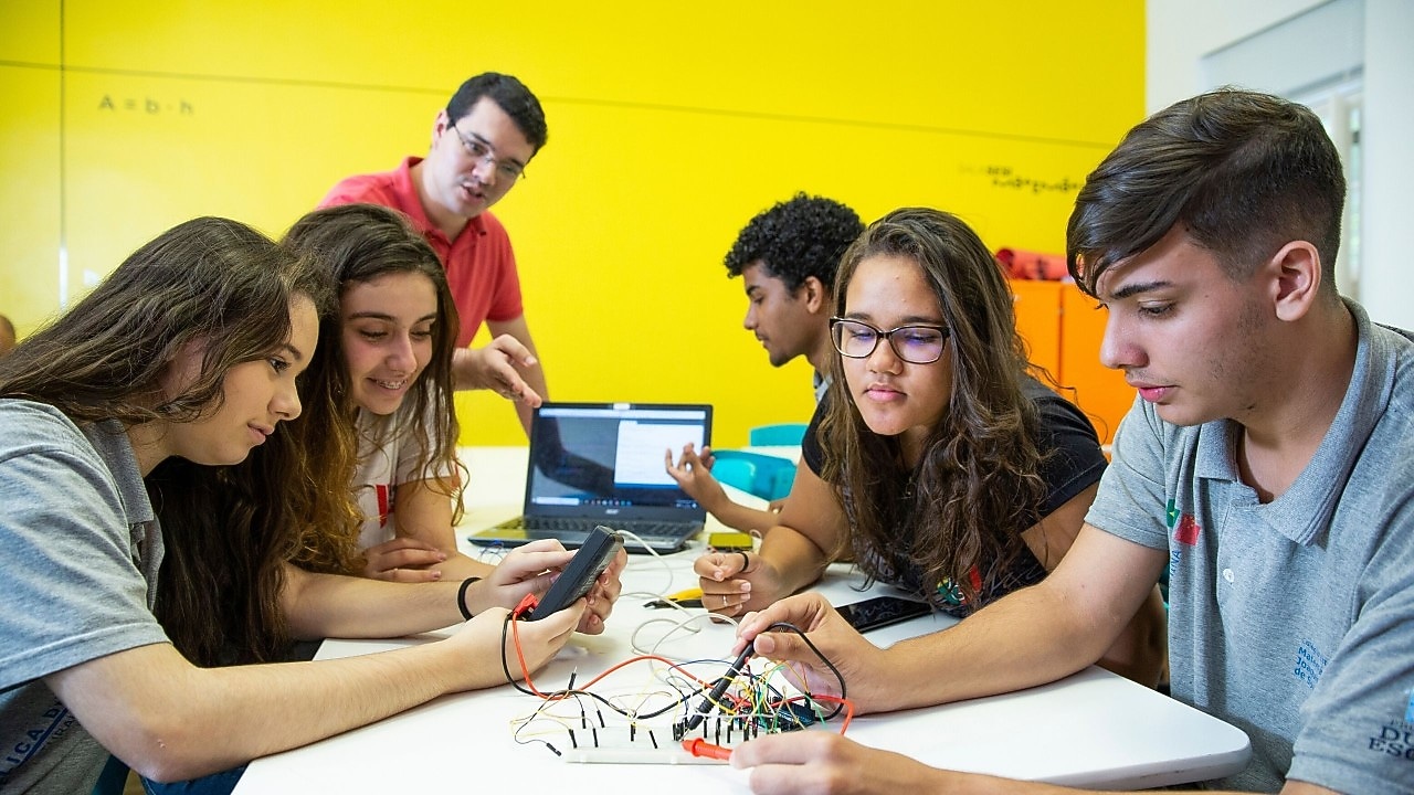 Na foto, vemos quatro alunos observando materiais do projeto. Ao fundo, um aluno de perfil ao lado de um computador e o professor explicando o projeto.