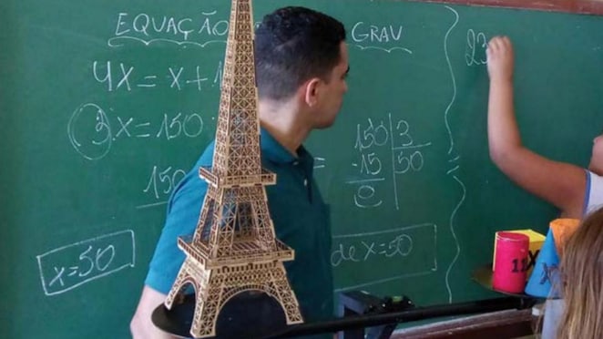Imagem de uma balança com uma réplica da Torre Eiffel do lado esquerdo e elementos geométricos coloridos do lado direito. Ao fundo, vemos um professor observando o estudante elaborando um cálculo no quadro negro.