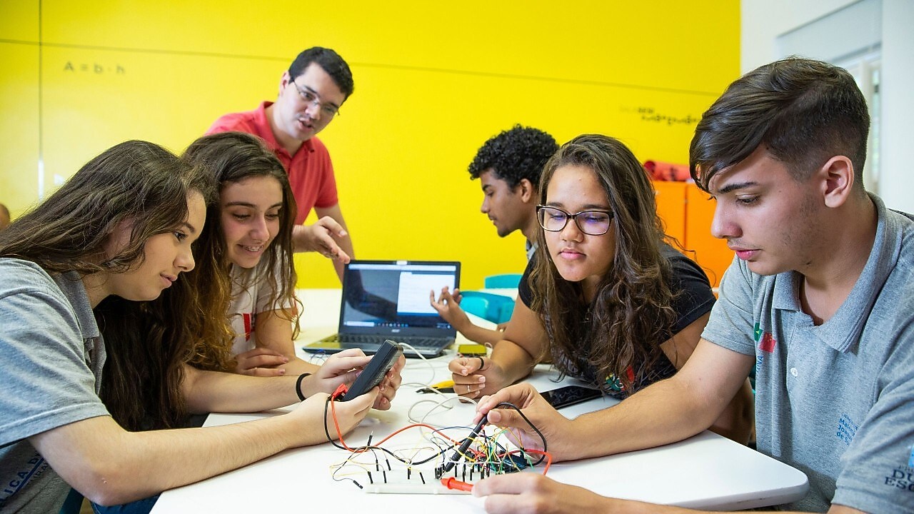 Na foto, vemos quatro alunos observando materiais do projeto. Ao fundo, um aluno de perfil ao lado de um computador e o professor explicando o projeto.