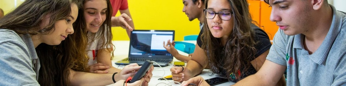 Na foto, vemos quatro alunos observando materiais do projeto. Ao fundo, um aluno de perfil ao lado de um computador e o professor explicando o projeto.