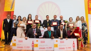 16 pessoas no palco posando para a foto. As pessoas formam duas fileiras no palco. Em pé atrás com há alguns vencedores com seus prêmio, flores e um cheque. Além de duas pessoas da Shell e o presidente, André Araujo. Embaixo, há 4 pessoas agachados com seus cheques e prêmios. Ao fundo vemos painel amarelado e a concha da Shell.