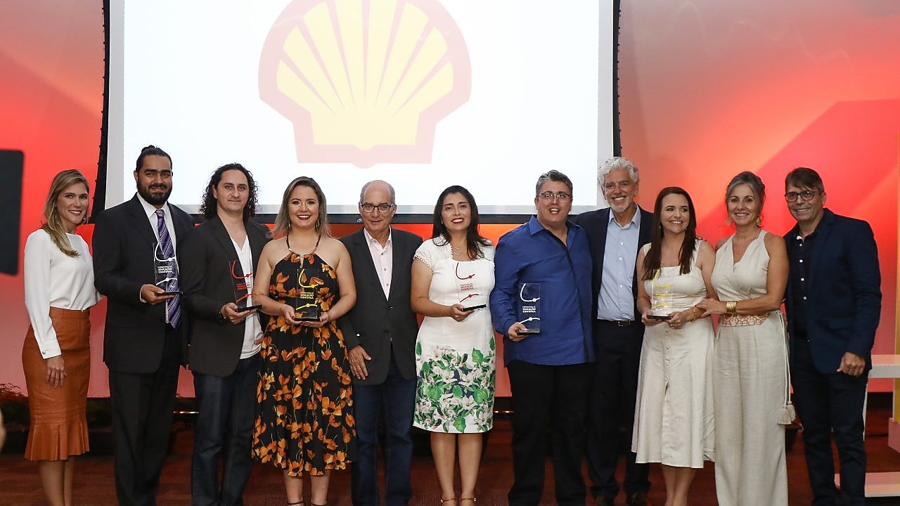 11 pessoas em pé posando para a foto. Dentre eles há 5 vencedores do Prêmio Shell de Educação Científica do Espírito Santo com seu trofeu, a apresentadora do prêmio e alguns dos avaliadores. Ao fundo temos uma luz avermelhada e um painel com a logo da concha.