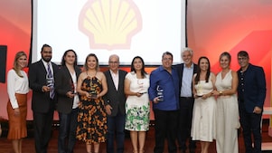 11 pessoas em pé posando para a foto. Dentre eles há 5 vencedores do Prêmio Shell de Educação Científica do Espírito Santo com seu trofeu, a apresentadora do prêmio e alguns dos avaliadores. Ao fundo temos uma luz avermelhada e um painel com a logo da concha.
