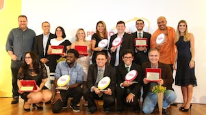 14 pessoas no palco posando para a foto. As pessoas formam duas fileiras no palco. Em pé há alguns vencedores com seus prêmios, além do André Araujo (presidente da Shell) e a realizadora do evento. Embaixo, há 5 vencedores agachados com seus prêmios.