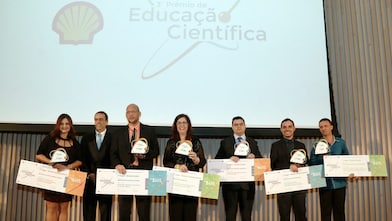 6 vencedores do Prêmio de Educação Científica no palco, segurando seus prêmios e cheques. Da direita para a esquerda, se encontra o presidente da Shell, André Araujo. Ao fundo há um telão com a logo da Shell e escrito terceiro prêmio de educação científica
