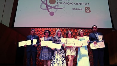 6 vencedores no palco do Prêmio de Educação Científica, segurando seus cheques e prêmios. Ao fundo há um telão com a logo da BG e do prêmio.