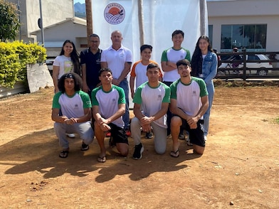 Grupo de dez pessoas, entre adolescentes e adultos, posa para a câmera em uma área externa ensolarada. Quatro adolescentes estão ajoelhados na frente e os demais permanecem em pé ao fundo. A maioria veste camisetas brancas com mangas verdes, enquanto atrás do grupo há um banner com o logotipo "Agrosol Coffee", além de construções, árvores e montanhas ao fundo, indicando uma visita ou atividade em uma propriedade ligada à produção de café.