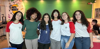 Grupo de seis adolescentes sorri para a câmera em um ambiente interno de criação e tecnologia. Cinco delas vestem camisetas escolares brancas com mangas vermelhas, enquanto uma usa uma blusa verde e outra uma camiseta esportiva azul. Ao fundo, há uma parede verde e um mural colorido com a inscrição "Arte Maker", além de mesas e equipamentos que sugerem um espaço voltado para atividades criativas e colaborativas.