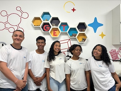 Grupo de oito jovens posa sorrindo para a câmera em uma sala de aula. Dois deles seguram um cartaz com o título "Círculo de Conexões", contendo anotações e linhas que conectam diferentes temas. Os demais estão posicionados ao redor do cartaz. Ao fundo, há um quadro branco, um relógio digital na parede e mesas, indicando uma atividade colaborativa ou apresentação escolar.