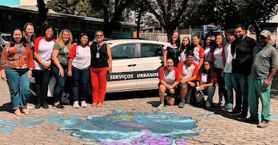 Grupo de adolescentes e adultos posa ao lado de um veículo identificado com a inscrição "Serviços Urbanos" em uma área externa arborizada. À frente do grupo, há um desenho colorido feito no chão, sugerindo uma atividade artística ou de conscientização. Todos estão voltados para a câmera, em um momento de registro coletivo relacionado a uma ação comunitária ou educativa.