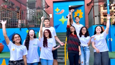 Grupo de oito adolescentes sorri e acena para a câmera em frente à entrada de um prédio colorido. Alguns levantam os braços em comemoração, enquanto estão posicionados ao redor de uma escada com corrimão amarelo. Ao fundo, a fachada azul tem ilustrações em formas geométricas coloridas, cercada por paredes de tijolos aparentes e vasos de plantas nas laterais.