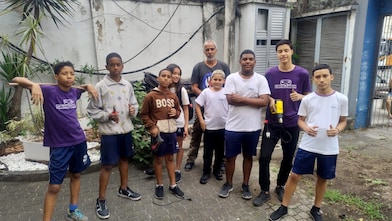 Grupo de nove adolescentes e um adulto posam ao ar livre em um pátio, olhando para a câmera. Alguns seguram ferramentas de jardinagem e mudas de plantas, enquanto outros fazem sinal de positivo com as mãos. Ao fundo, há um prédio com paredes claras, janelas com venezianas, vegetação e canteiros, sugerindo uma atividade de plantio ou manutenção de área verde.