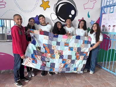 Grupo de sete adolescentes sorri para a câmera enquanto segura uma colcha de retalhos colorida estendida à frente. Eles estão em um espaço interno com parede decorada por ilustrações coloridas, incluindo um astronauta estilizado e estrelas. A cena transmite um momento de celebração e colaboração em torno da peça produzida.