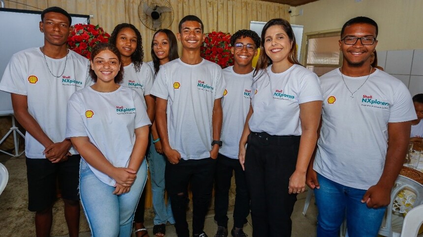 A imagem mostra os alunos do grupo e a professora, vestindo camisetas brancas com o mesmo logotipo na área do peito esquerdo, que inclui um elemento laranja e o texto "Nxplorer".