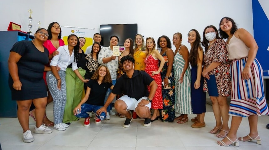 A imagem mostra o grupo de alunos reunidos com os professores em um ambiente interno, de paredes brancas adornadas com certificados emoldurados e uma prateleira exibindo troféus, sugerindo um ambiente de conquista ou reconhecimento.