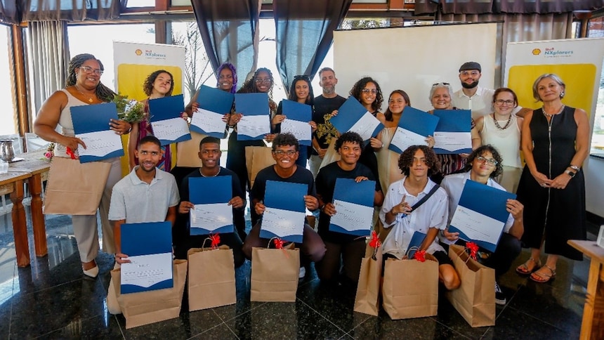 A imagem retrata o grupo de alunos e professores em um ambiente interno, uma sala com grandes janelas ou portas de vidro ao fundo. Cada aluno está segurando um certificado azul e um buquê de flores, e há sacolas de presente marrons aos seus pés.