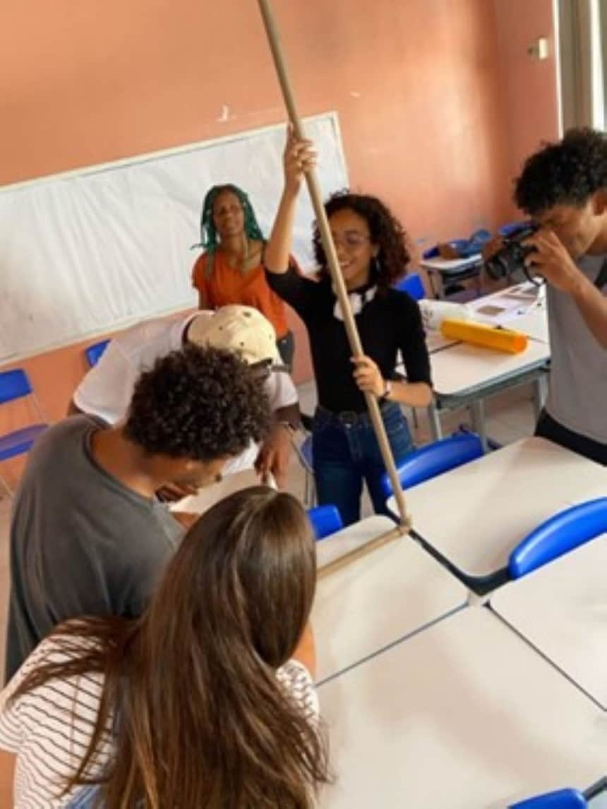 A imagem mostra os alunos do grupo ao redor de uma mesa em um ambiente de sala de aula, com uma das pessoas segurando o que parece ser uma vara ou poste longo. Há um quadro branco ao fundo, mas nenhum texto visível.