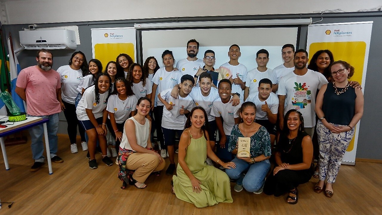A imagem mostra o grupo de alunos e professores um ambiente interno que parece ser uma sala de aula. O local está bem iluminado e, no fundo da sala, há banners pendurados na parede.