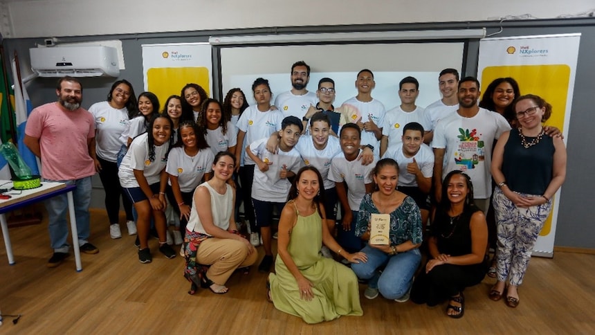 A imagem mostra o grupo de alunos e professores um ambiente interno que parece ser uma sala de aula. O local está bem iluminado e, no fundo da sala, há banners pendurados na parede.