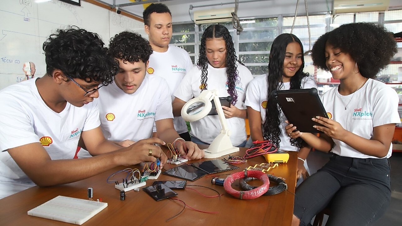 A imagem mostra os alunos ao redor de uma mesa, envolvidos no que parece ser uma oficina de eletrônica. Na mesa estão espalhados vários componentes, como fios, placas de circuito e ferramentas. Uma aluna está segurando um tablet, enquanto outro está trabalhando na montagem ou reparo de um dispositivo eletrônico com um ferro de solda. Um quadro branco com algumas anotações nele é visível ao fundo, mas o texto não é legível.