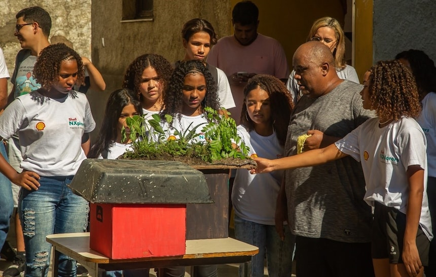 A imagem mostra o grupo de alunos, em pé ao ar livre à luz do dia. Eles estão segurando um modelo de uma casa que tem plantas verdes crescendo em cima dela, simbolizando talvez um telhado verde ou um conceito de habitação ecológica. O modelo é colocado em uma placa de madeira para suporte. Os alunos estão vestindo camisetas brancas com logotipos nelas.