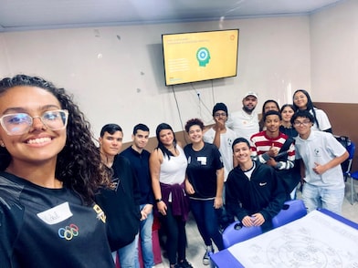Selfie de um grupo de cerca de 13 pessoas em uma sala de aula ou treinamento. Em primeiro plano, à esquerda, uma mulher de cabelos cacheados e óculos sorri para a câmera enquanto tira a foto. Ao fundo, os demais participantes estão reunidos em pé e sentados, sorrindo e posando juntos. Na parede, há uma televisão exibindo um slide com o título "Shell promove programa de educação digital", acompanhado de um ícone ilustrativo. No canto inferior direito, uma mesa azul exibe folhas de papel com um diagrama impresso. O ambiente é simples, com paredes claras, cadeiras coloridas e clima descontraído de atividade em grupo.