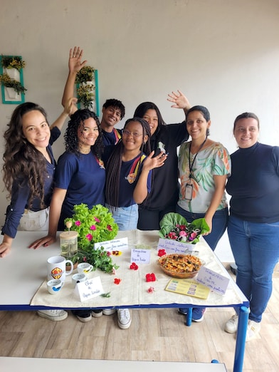 Grupo de sete pessoas posa sorrindo atrás de uma mesa com plantas, flores e alimentos, em um ambiente interno. Algumas acenam para a câmera, criando um clima descontraído. Sobre a mesa há vasos com flores e hortaliças, uma torta, copos, um pote de vidro e pequenos cartazes escritos à mão com informações sobre os itens expostos. Ao fundo, uma parede clara e elementos decorativos com folhagens completam o ambiente, sugerindo uma atividade ou feira voltada à jardinagem, sustentabilidade ou alimentação.