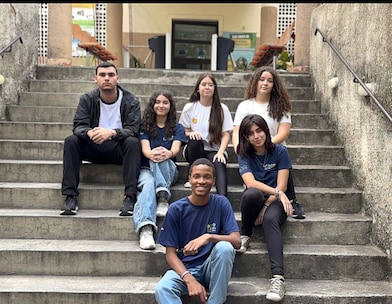 Seis jovens posam sentados em uma escadaria de concreto ao ar livre, olhando para a câmera. Um rapaz está sentado no degrau mais baixo, sorrindo, enquanto os outros cinco ocupam os degraus superiores. O grupo veste roupas casuais, com camisetas em tons de azul, branco e preto, e transmite um clima descontraído. Ao fundo, há a entrada de um prédio, com colunas, corrimãos e um jardim parcialmente visível.