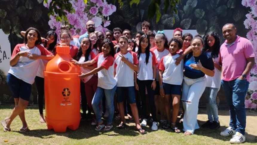 A imagem mostra os alunos do grupo junto com os professores em um ambiente ao ar livre. Eles estão em pé, sorrindo, e estão posando com uma grande garrafa com o símbolo de reciclagem estampado.