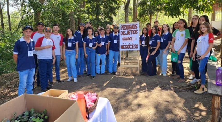 A imagem mostra o grupo de alunos em pé ao ar livre, com uma faixa que diz "FEIRAS DE TROCA DE OBJETOS E NEGÓCIOS". O cenário parece ser um parque ou um espaço verde semelhante, pois há árvores e plantas ao fundo. Há também uma caixa com o que parecem ser plantas ou flores no canto inferior esquerdo da imagem.